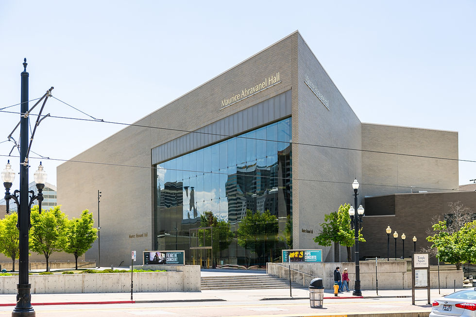 Modern building of Maurice Abravanel Hall with reflective glass facade. Trees and signs for concerts in front, clear sky above.