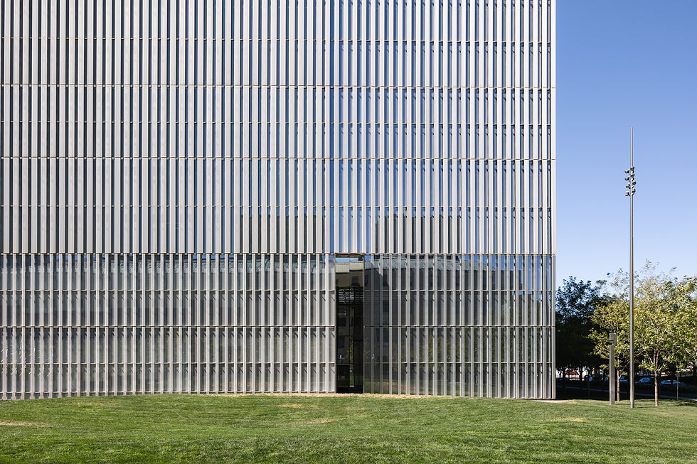 Modern building with vertical glass panels, reflecting blue sky. Green lawn in foreground and trees on the right. Calm urban setting.