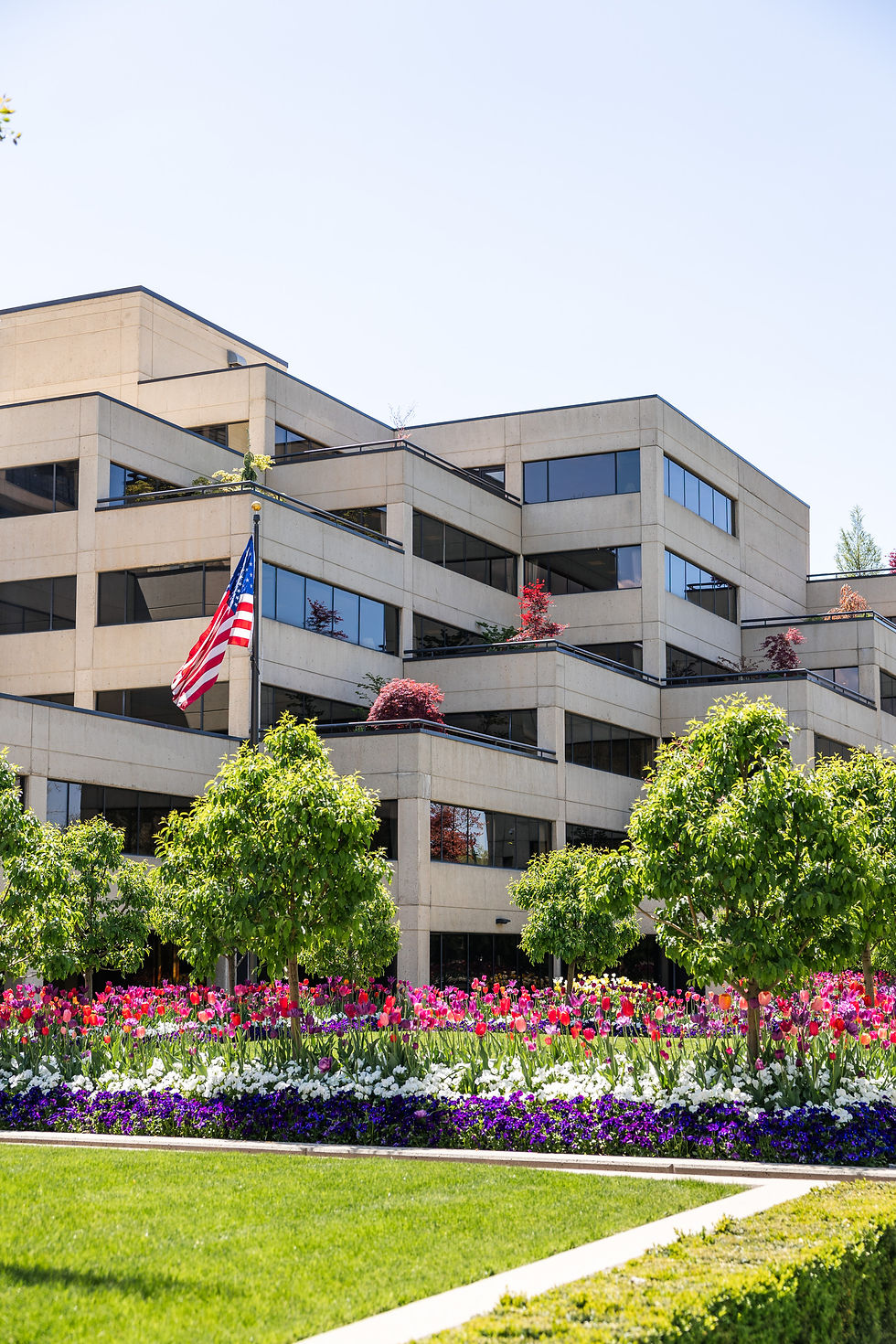 Modern building with beige exterior, large windows, and an American flag. Lush garden with green trees, colorful flowers, and vibrant greenery.