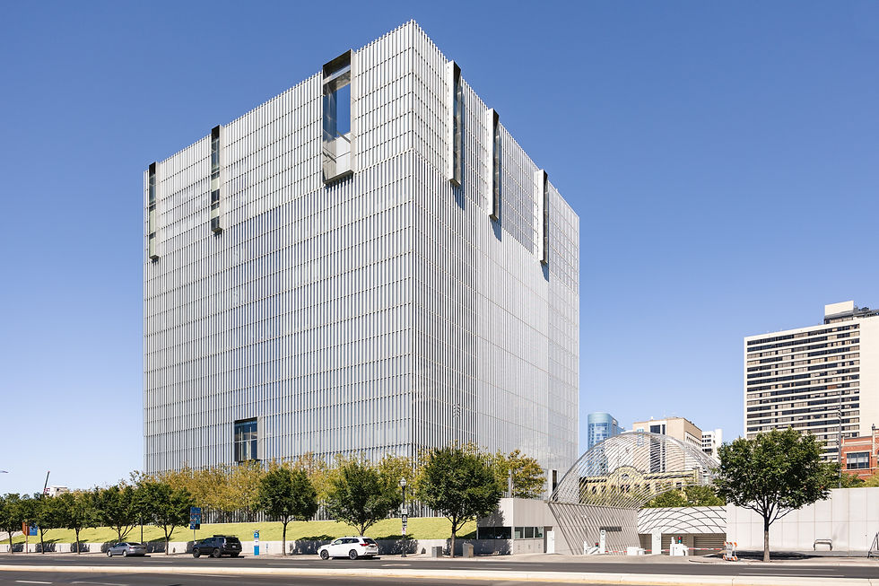 Modern glass and metal building under clear blue sky. Trees line the street with parked cars. Urban setting with skyscrapers in the background.