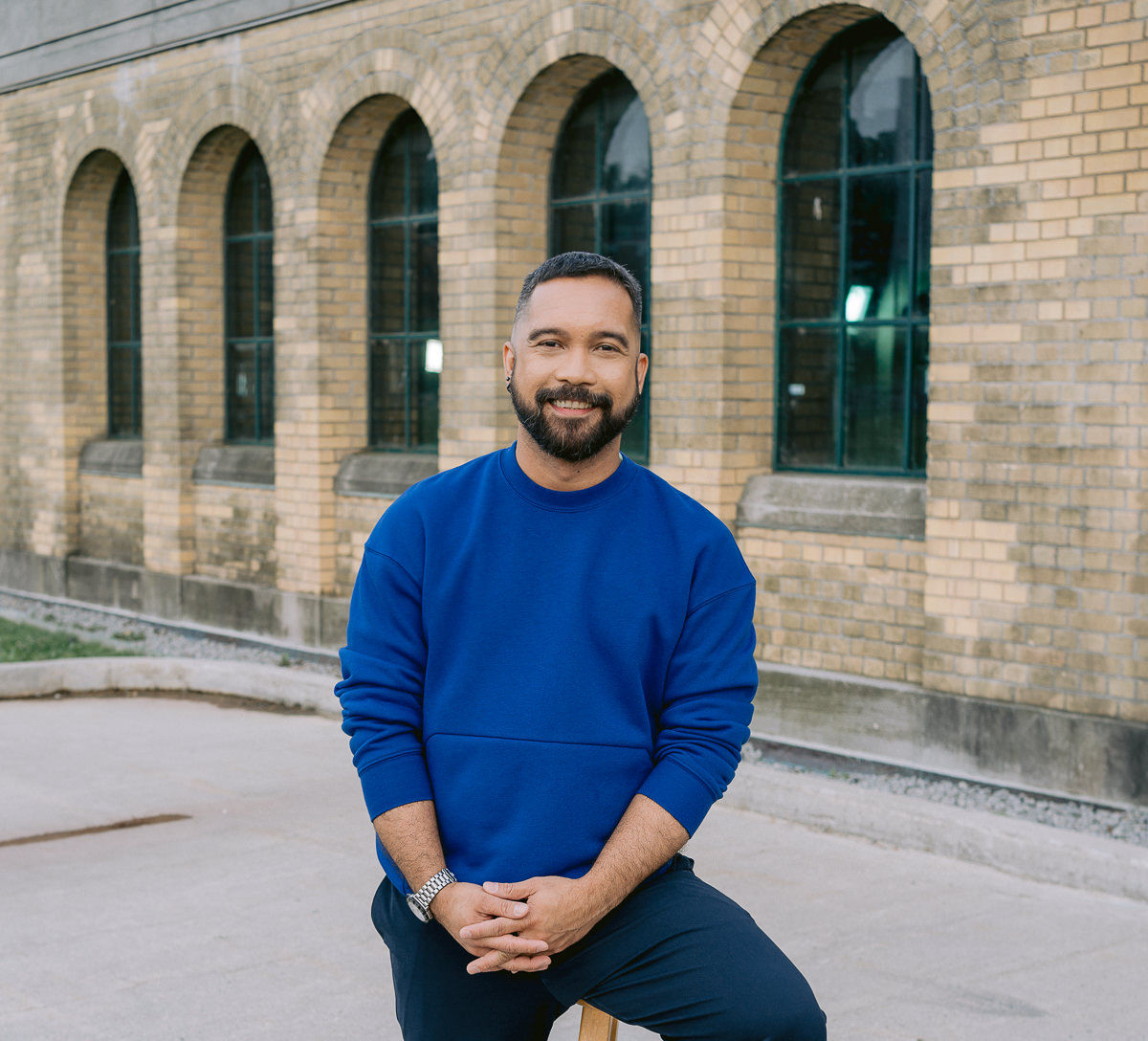 Michael, in blue, hands together, sitting on a stool in front of a brick building. 