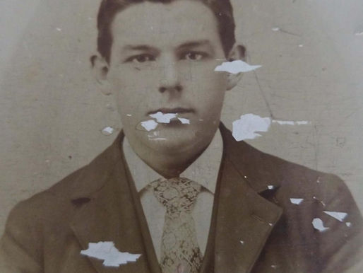 Vintage portrait of a young man in a suit and patterned tie, with a neutral expression. The image is worn and faded with visible damage.