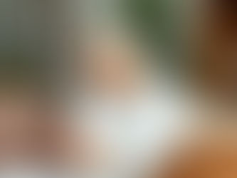 Elderly woman with glasses sits on a beige sofa using a smartphone. Cozy room with plants and shelves. Calm and focused expression.