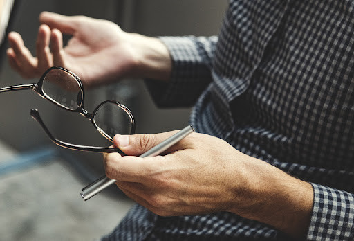 Man with his hands out, one of which is holding a pair of glasses and a pen.