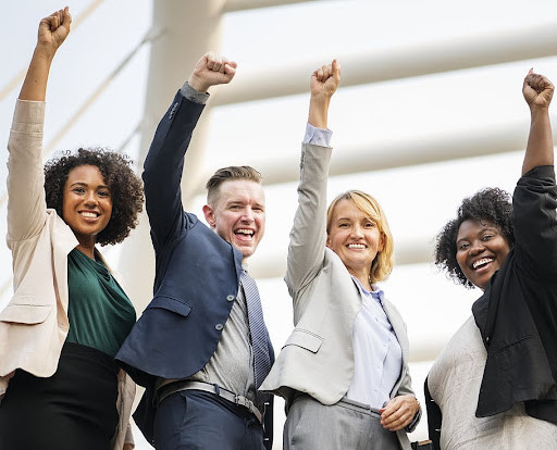 Four people in business attire cheering together. One is a white man with a slicked back hair, another is a white woman with wavy blond hair, and the other two are black women with voluminous curly dark hair.