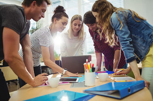 A team of people gathered around table and discussing a project. Binders, pens, cups, and various electronics are littered across the table.