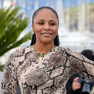 Smiling woman in snake-pattern dress stands outdoors with palm trees and sea in the background. Photographers are visible.