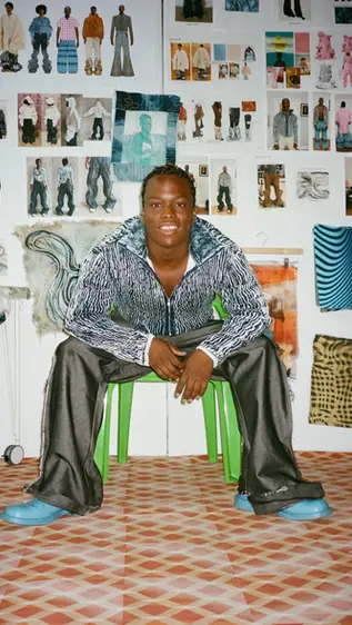 Man in patterned jacket sitting on green chair, smiling. Background full of fashion sketches and fabrics. Floor has geometric tiles.