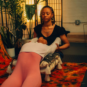 Two people engage in a calming, supportive pose indoors, surrounded by plants and soft lighting. Warm colors create a serene mood.