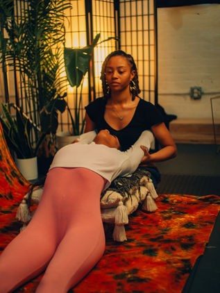 Two people engage in a calming, supportive pose indoors, surrounded by plants and soft lighting. Warm colors create a serene mood.