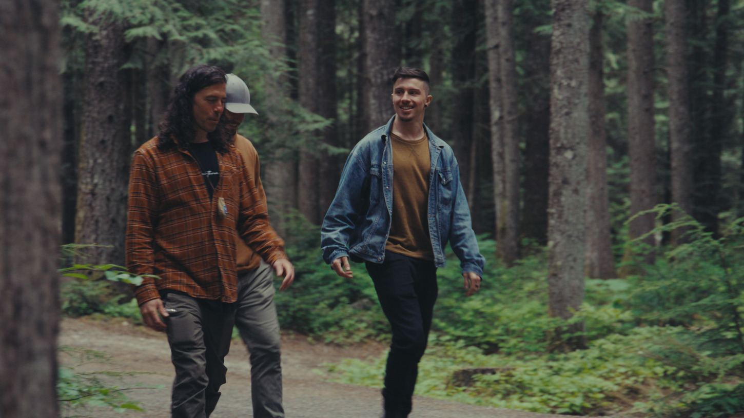 Two men walk along a forest path. One wears a brown plaid shirt and a cap, the other a denim jacket. They appear relaxed and are surrounded by tall trees.