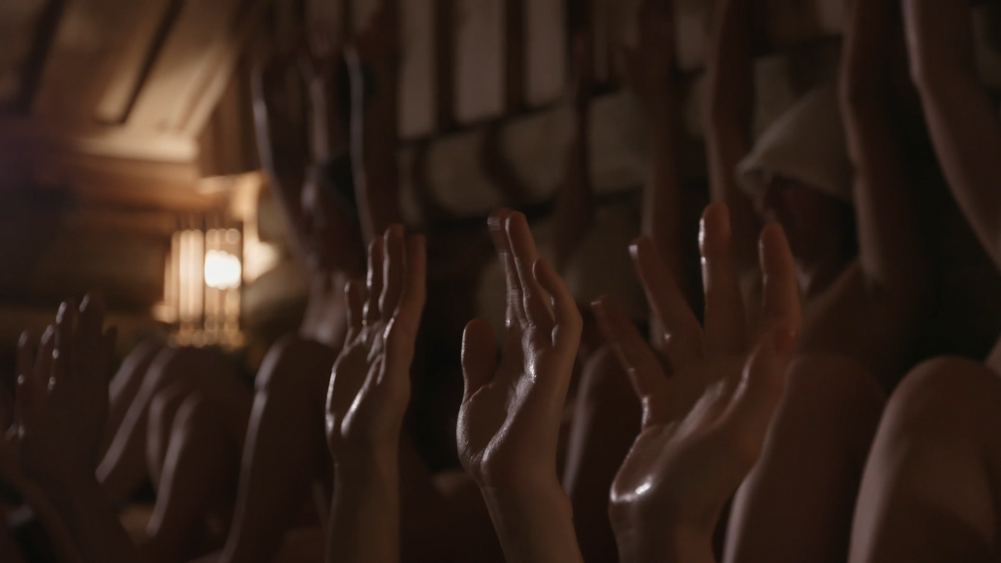 Hands raised towards a dimly lit ceiling in a sauna, with a soft glow from a lamp creating a warm, tranquil atmosphere.