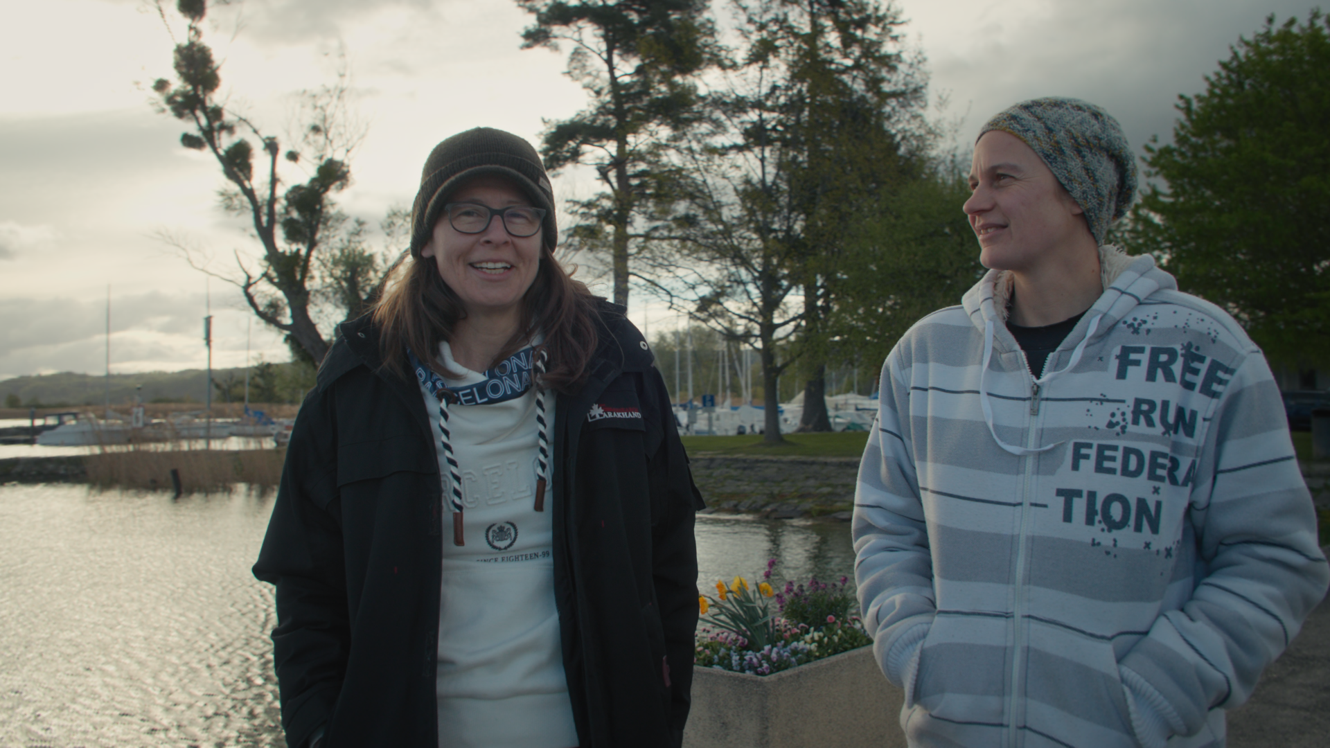 Two individuals stand by a calm waterfront in Erlach, Switzerland, smiling and wearing casual, warm clothing. Trees and boats are visible in the background, under a cloudy sky.