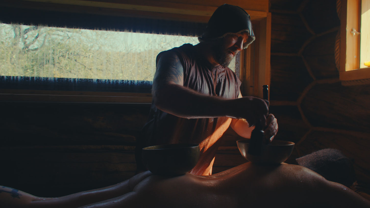 A man massages a nude woman's back in a dim, rustic room on Sauna Channel. Soft natural light filters through a window, creating a warm, serene, and calming atmosphere.