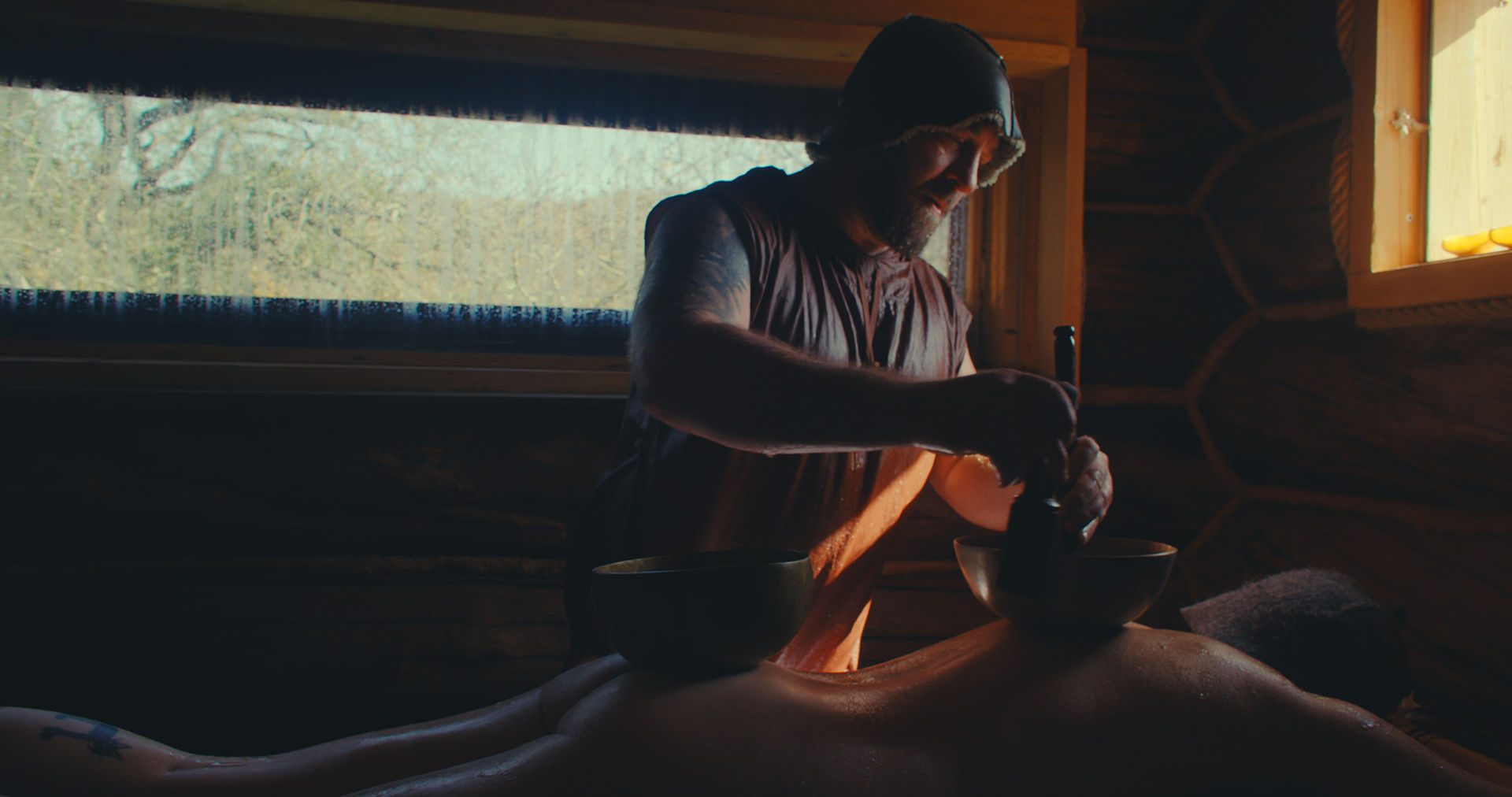 גבר מעסה את גבה של אישה עירומה בחדר כפרי ואפלולי בערוץ הסאונה. אור טבעי רך מסתנן דרך החלון ויוצר אווירה חמימה, שלווה ומרגיעה.