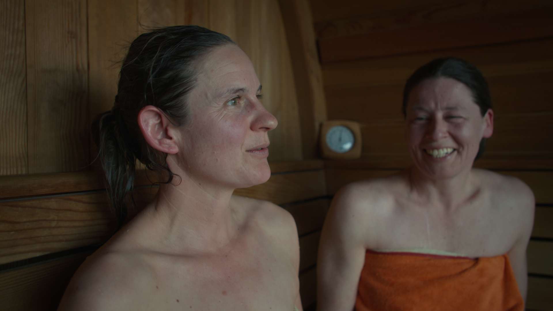 Two women sit in a wooden sauna, both with relaxed expressions. One wears an orange towel. A round thermometer is visible on the wall behind them.