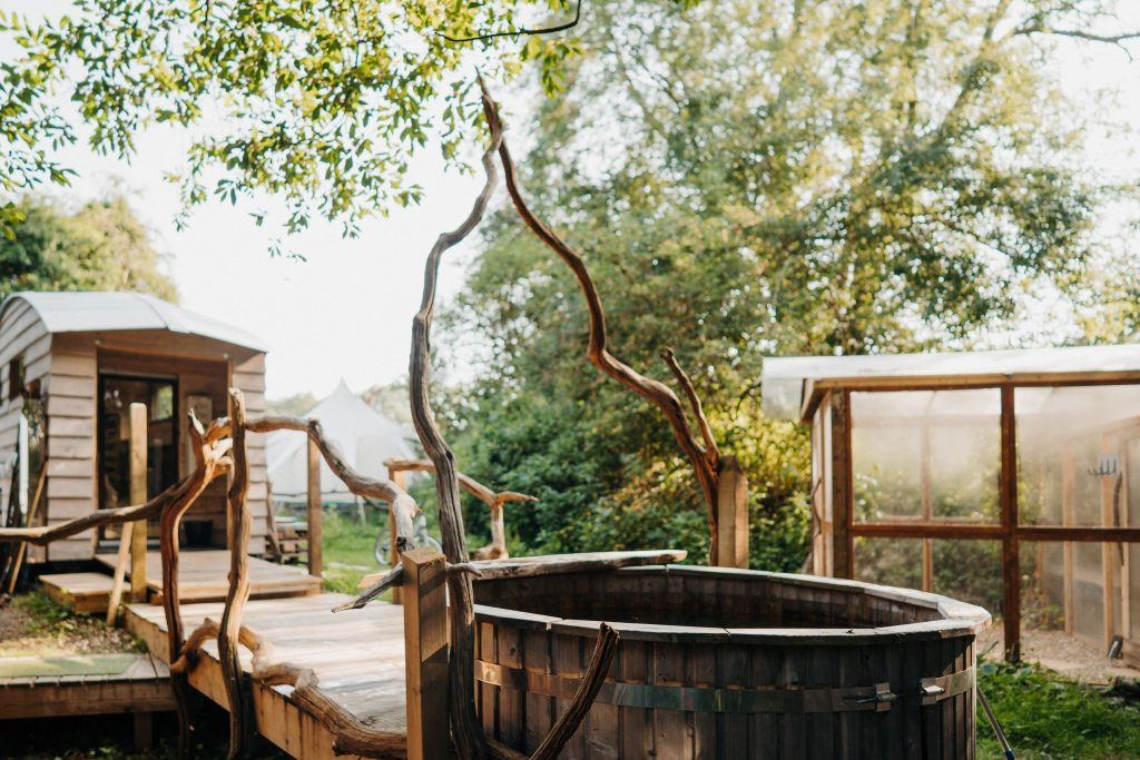 An outdoor hot tub with earthy tones next to trees and a wooden bridge with twisting wood branch railings at Wild Spa Wowo in Sussex, England on Sauna Channel