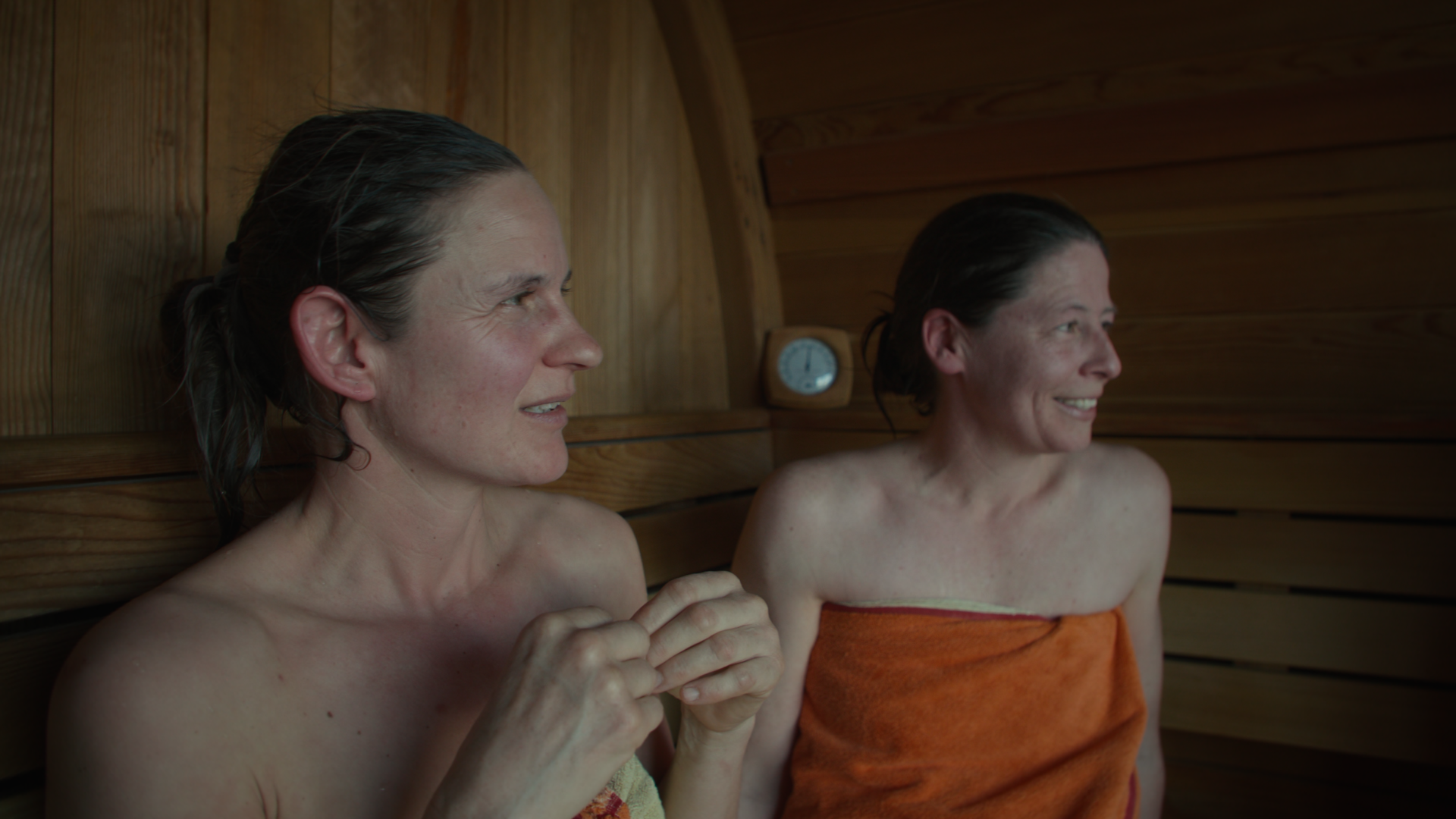 Helena and Sibylle sit in a wooden sauna in Switzerland wrapped in towels, smiling and looking to the right. The atmosphere is relaxed and warm, with soft lighting.