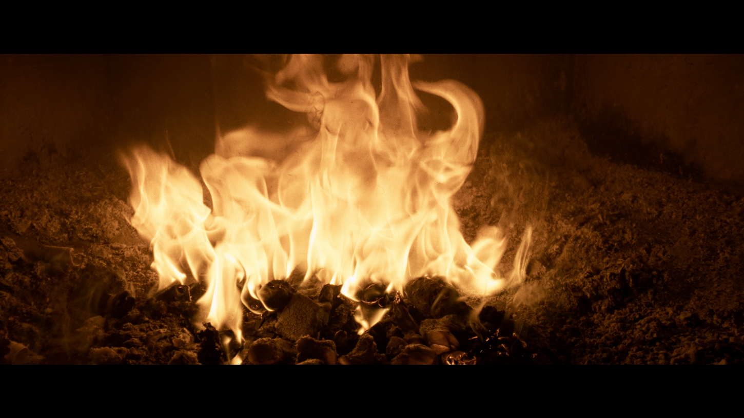 Intense close-up of vibrant yellow-orange flames dancing over glowing coals, creating a warm, dynamic, and fiery atmosphere in a dimly lit setting on Sauna Channel.