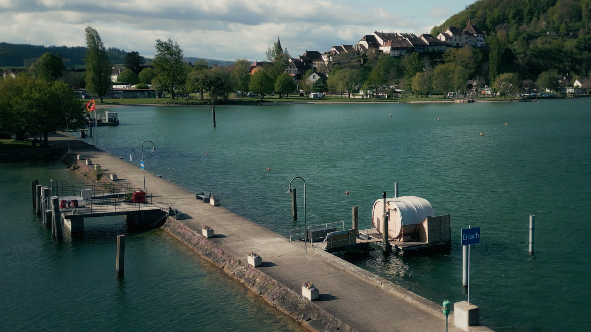 A serene lakeside scene featuring a dock extending into calm water, a small boat, and lush greenery in The Swiss Sauna Raft episode of Sauna Channel.