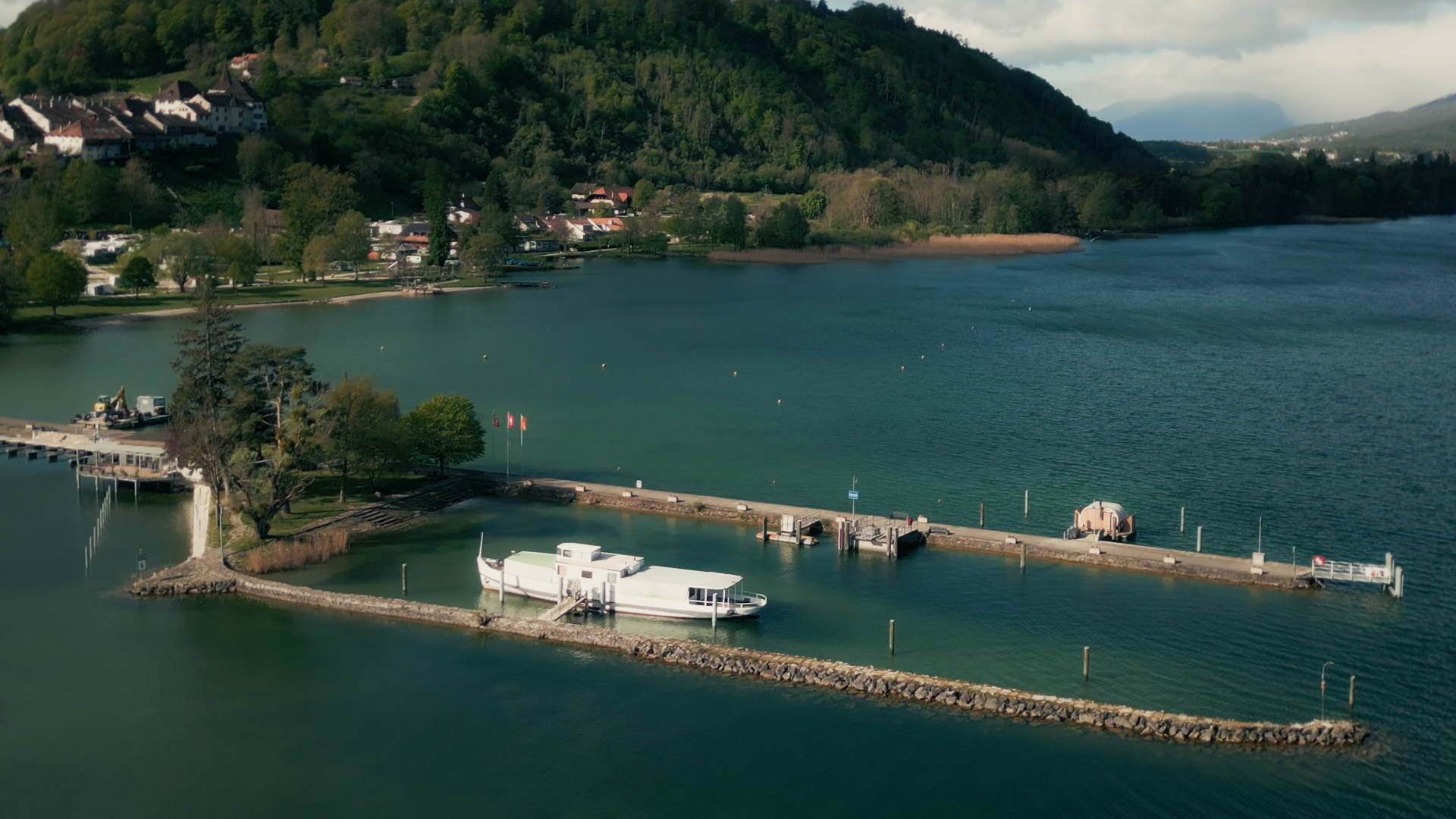 Aerial view on Sauna Channel of a tranquil serene Switzerland lakeside with boats docked at a pier, surrounded by lush green hills and a small community of houses.
