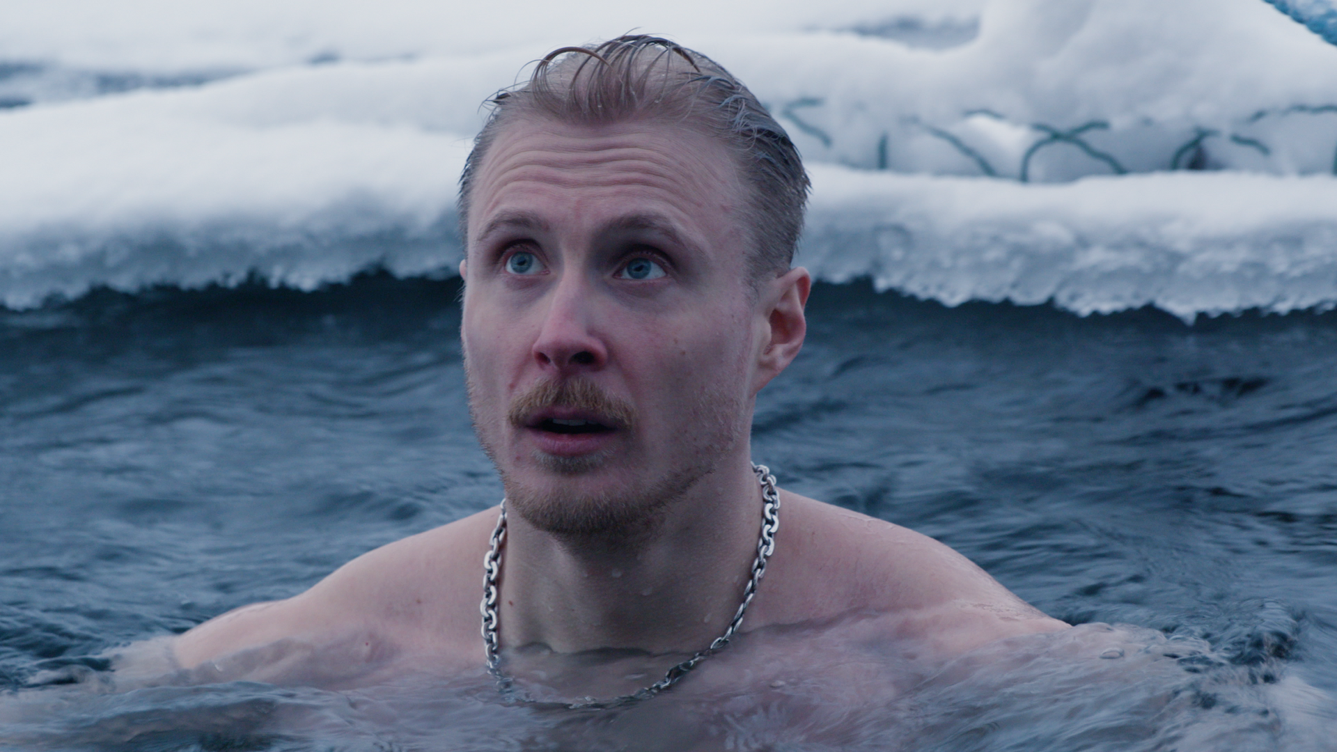 A man wearing a silver chain endures brutally cold ice river swimming in the heart of Finland's wilderness on the Sauna Channel episode: Kiilopää Smoke Sauna.
