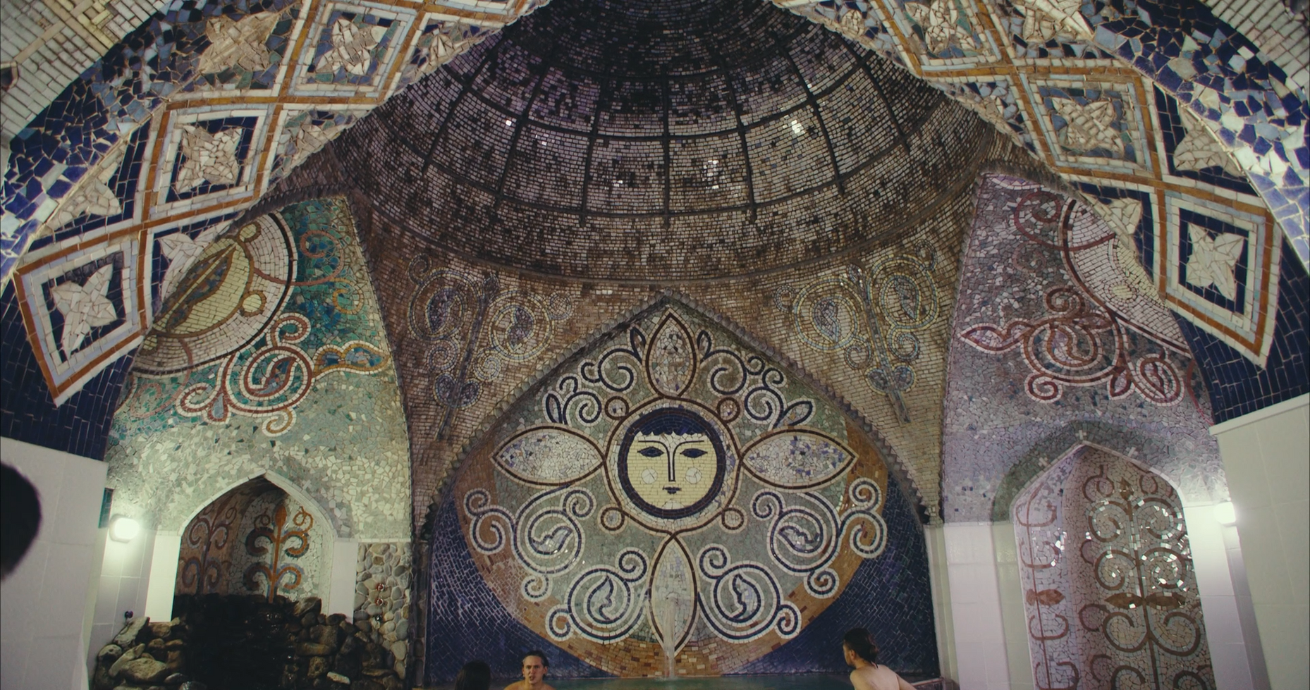 Intricate mosaic design featuring abstract patterns and a stylized face on a domed ceiling, evoking a sense of mystery and artistic grandeur.