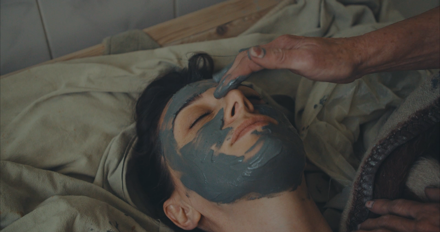 A person receives a gray face mask treatment, lying back peacefully with closed eyes. A hand gently applies the mask, conveying relaxation.