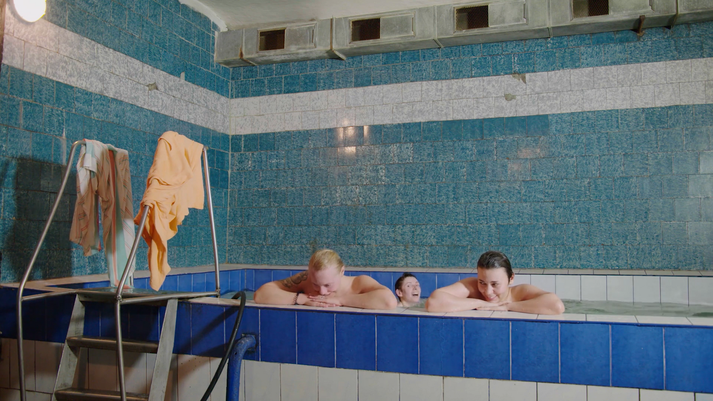 Three people in St. Petersburg relax in a small indoor pool with blue tiled walls, appearing content. Towels hang on a metal ladder nearby.