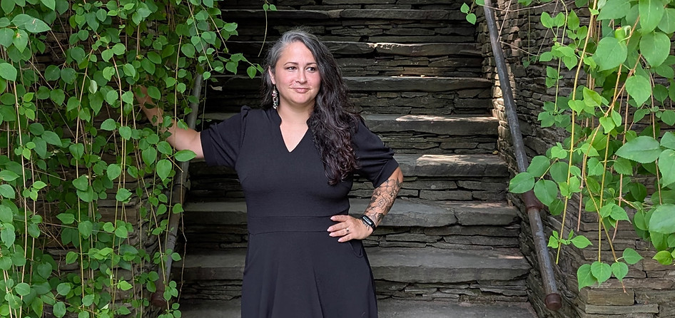 A woman photographer is standing in front of a stone staircase with a green foliage leaf backdrop while looking off into the distance. She has a black dress, tattoos, earrings and black and some grey hair while smiling.