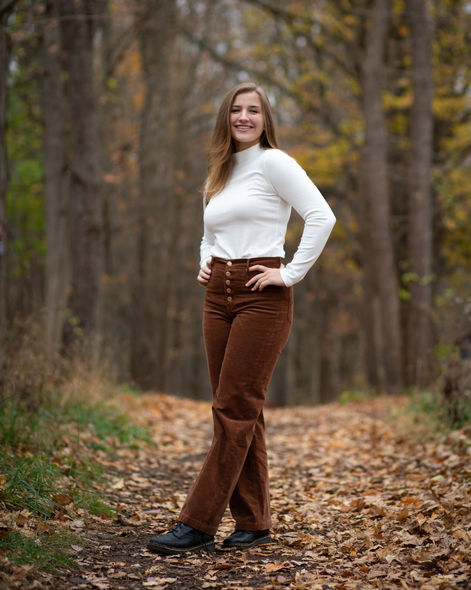 A teenager who is a high school senior is standing in the woods on a path with foliage in the fall time while posed and smiling.