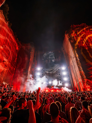 Crowd at a red-lit outdoor concert in a quarry, cheering before a giant projected stone face and bright stage lights.