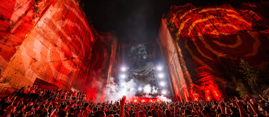 Crowd at a red-lit outdoor concert in a quarry, cheering before a giant projected stone face and bright stage lights.