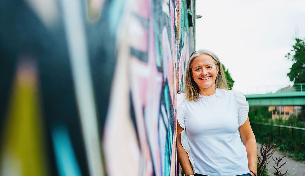 Jo is smiling at the camera, outdoors against a vibrant wall painted in colourful grafitti