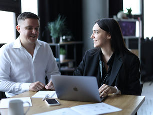 Deux personnes souriantes discutent autour d'un bureau avec un ordinateur portable et des documents. Arrière-plan de plante et étagère. Ambiance professionnelle.