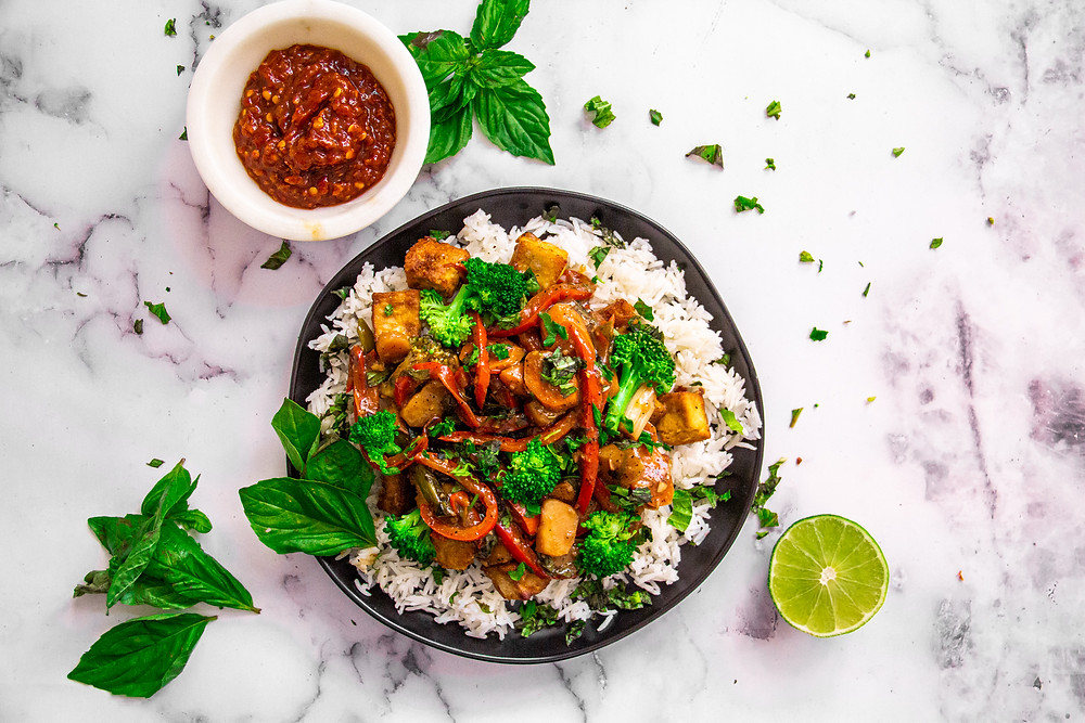 Black plate filled with rice, tofu, broccoli, red bell peppers, and basil.