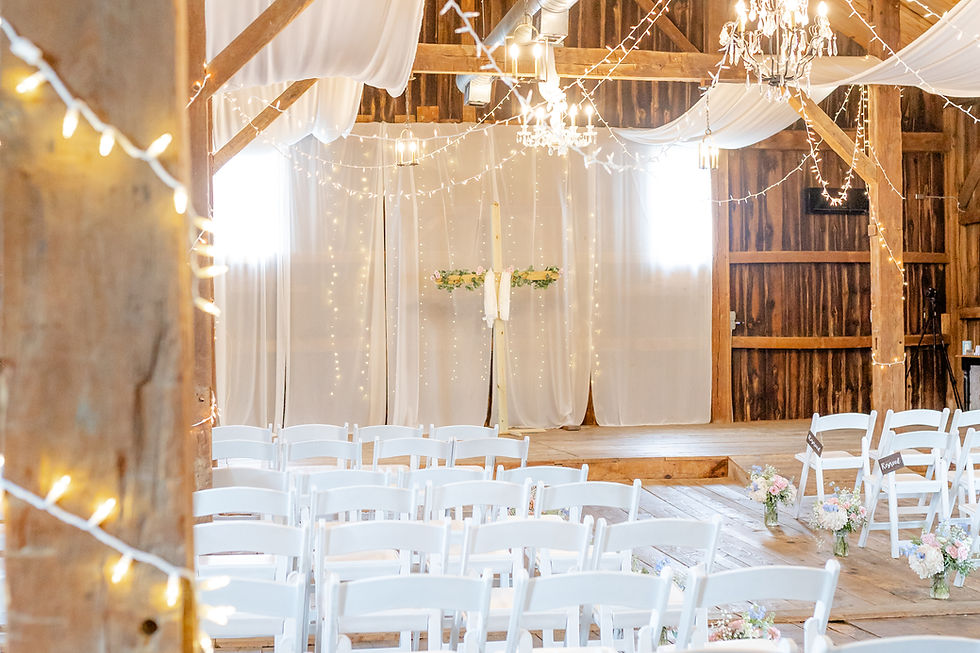 Rustic barn wedding ceremony setup with white chairs, floral arrangements, draped fabric, string lights, and reserved signs visible.