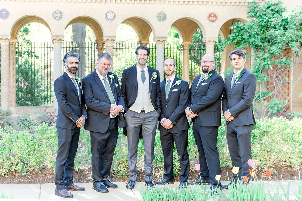 Six men in suits with boutonnieres stand smiling in a garden. Arched stone wall and colorful tulips in the background.