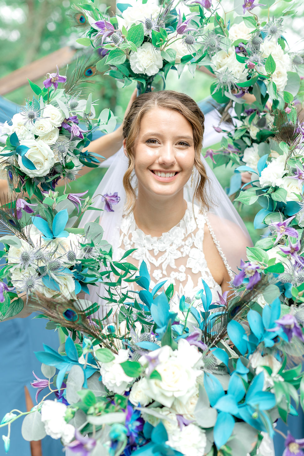 Smiling bride in lace dress framed by white, purple, and teal flowers and peacock feathers outdoors.