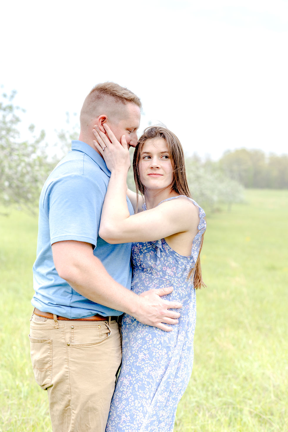 A couple embraces in a grassy field. The woman in a floral dress gazes pensively, while the man in a blue shirt holds her gently.
