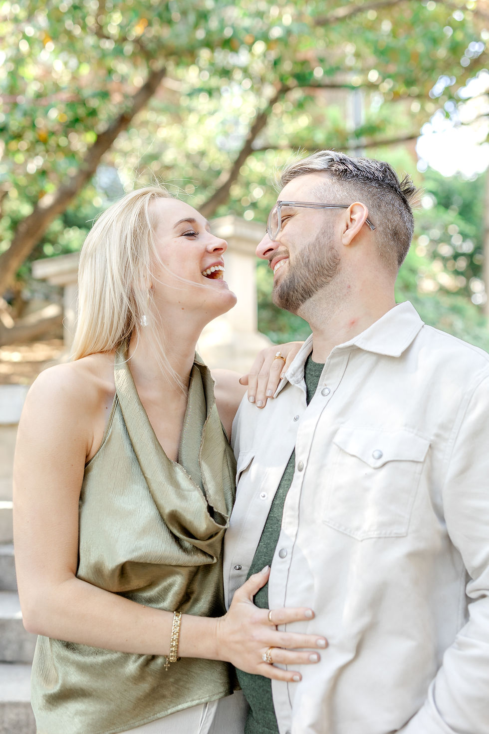 A smiling couple looks at each other in a park, with trees in the background. The woman wears a green top; the man wears glasses and a white shirt.