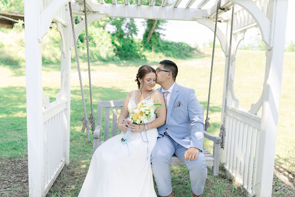 Bride and groom sit on a white garden swing; he kisses her forehead as she holds a bouquet, with green trees behind them.