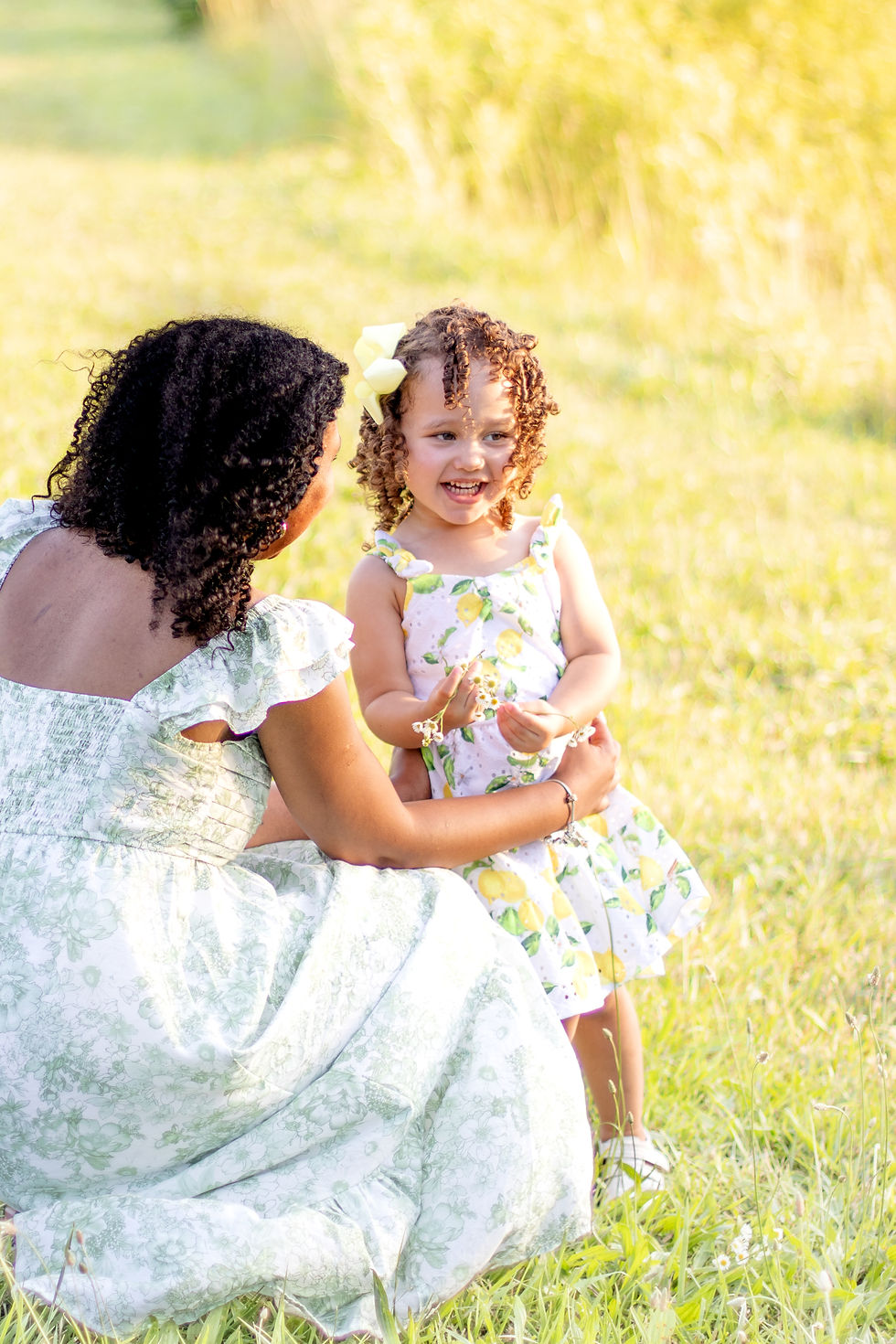 Woman and child in floral dresses enjoying a sunny field. The child holds flowers, smiling brightly. Bright, cheerful atmosphere.
