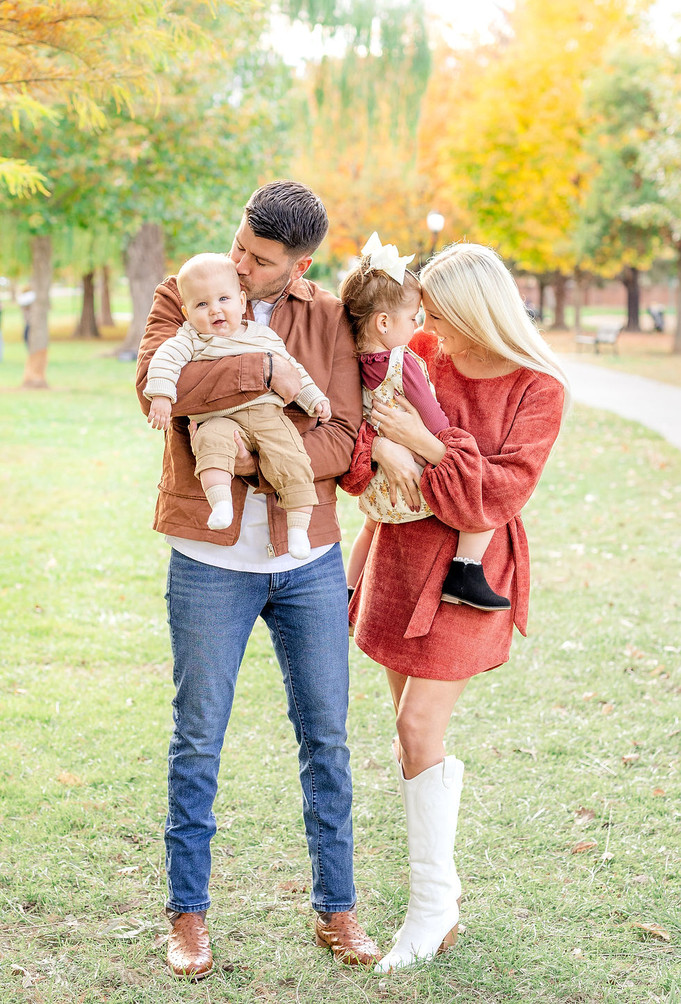 Family in a park: Dad kisses baby; mom holds toddler. Autumn leaves in background. Warm colors, joyful mood.