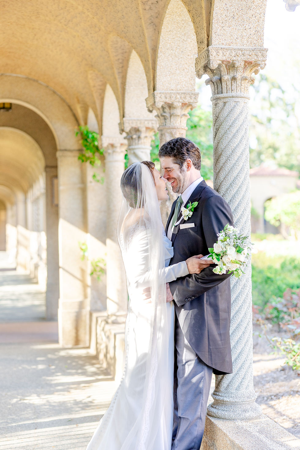 Bride and groom embrace and smile in a sunlit stone archway. Bride holds a white bouquet, wearing a veil. Romantic and joyful mood.