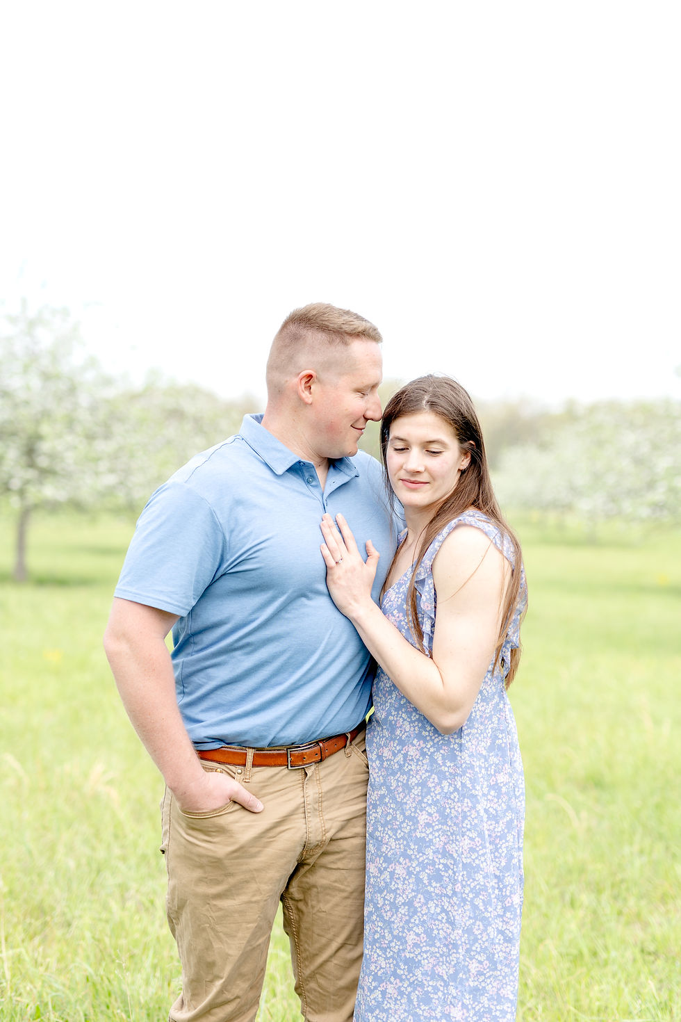 A couple stands in a field; the man in a blue shirt embraces the woman in a floral dress. The setting is a sunny, grassy landscape.