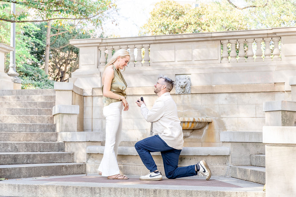 Man kneels with ring in hand proposing to woman on stone steps. Woman smiles warmly. Lush garden surrounds the elegant setting.