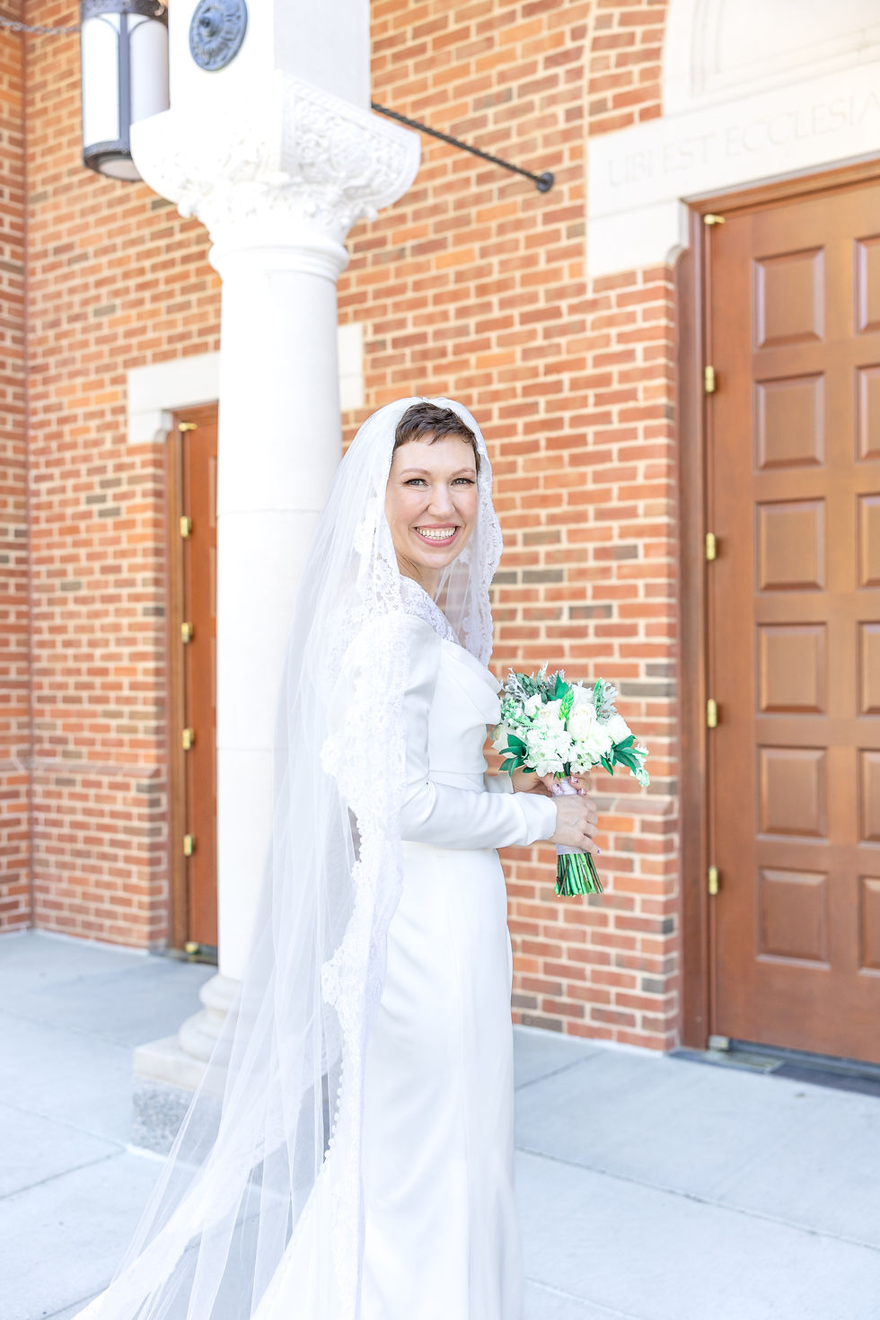A smiling bride in a white dress and lace veil holds a bouquet outside a brick building with wooden doors, under a column.