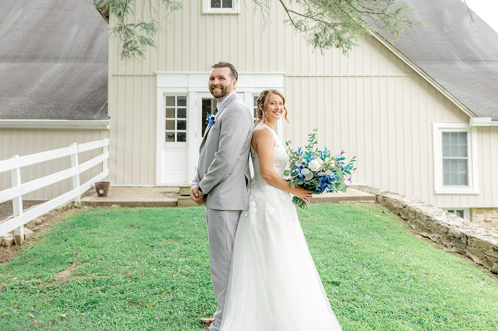 Smiling bride and groom stand back to back on a lawn in front of a beige barn, holding a blue-and-white wedding bouquet.
