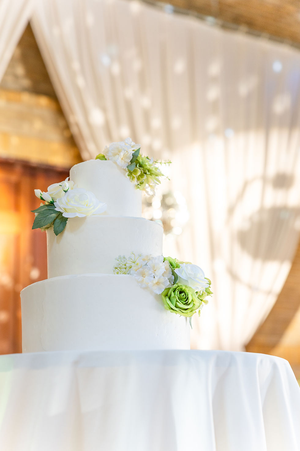 Three-tier white cake adorned with green and white flowers, set against a softly lit draped background. Elegant and celebratory mood.